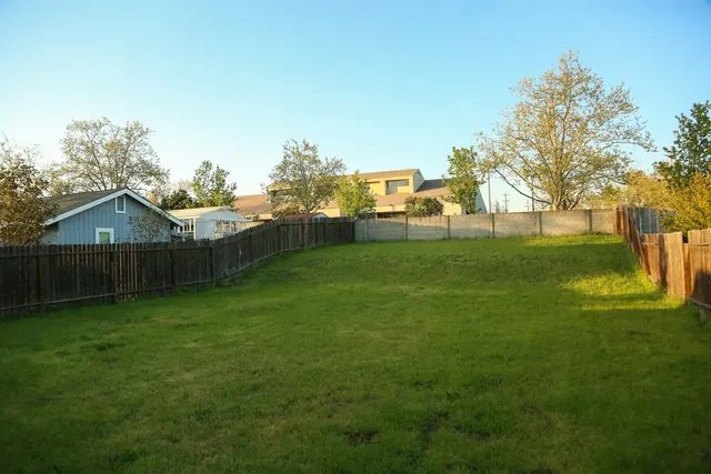 Before: a long empty lawn enclosed by dark timber fences with houses and trees beyond.