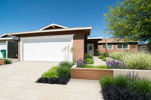 AI generated modern redesign of the same view: a wide curved concrete driveway leading to a double garage on a low brick-coloured house, replanted.