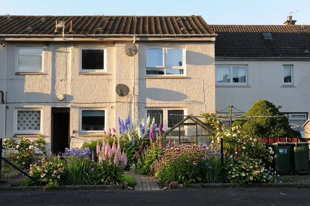 AI generated cottage redesign of the same view: the small front garden of a rendered terraced house with a bench, a small greenhouse and wheelie bins, replanted.