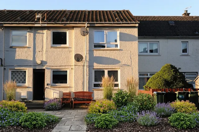 AI generated low maintenance redesign of the same view: the small front garden of a rendered terraced house with a bench, a small greenhouse and wheelie bins, replanted.