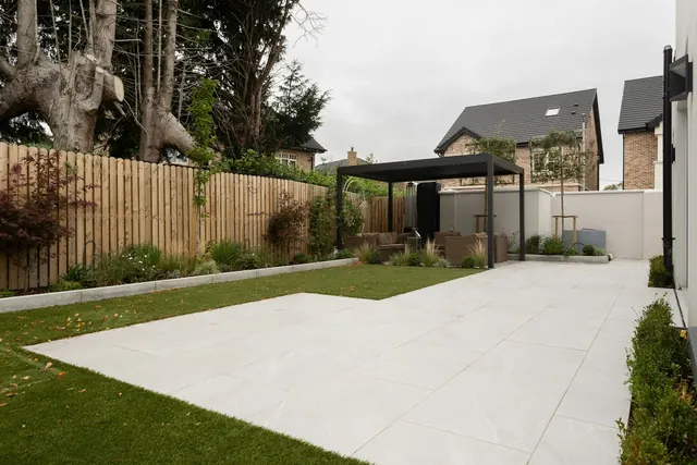 Before: a large pale porcelain slab patio beside a small lawn with a timber fence, a pergola and neighbouring houses.