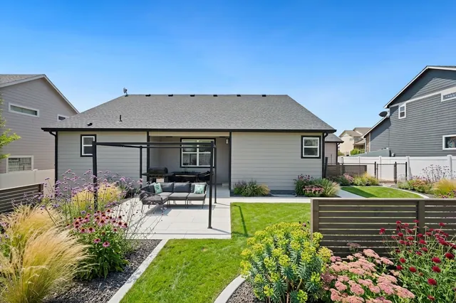 AI generated modern redesign of the same view: a back garden of mown lawn with a small concrete slab patio and metal pergola behind a single-storey house, replanted.