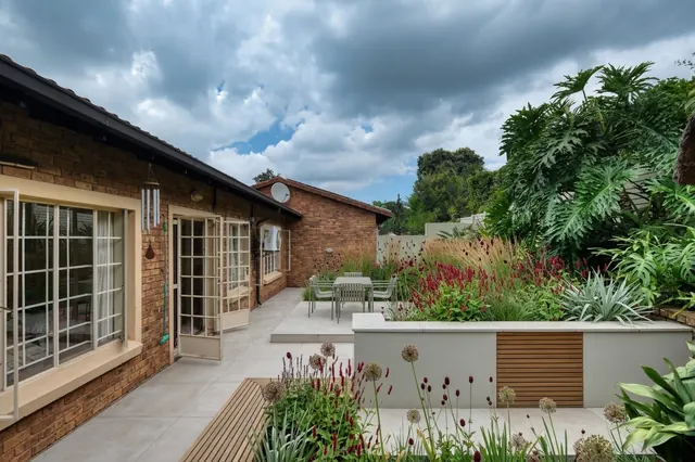 AI generated modern redesign of the same view: a narrow side garden of lawn and a brick paved patio running along a low brick wall beside a brick house, replanted.