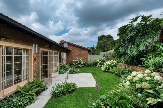 AI generated shade planting redesign of the same view: a narrow side garden of lawn and a brick paved patio running along a low brick wall beside a brick house, replanted.