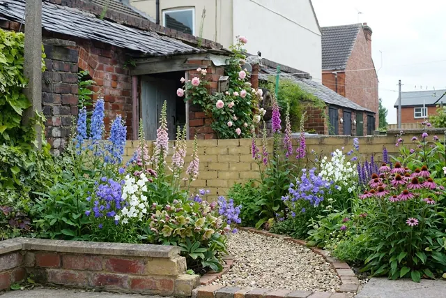 AI generated cottage redesign of the same view: a dilapidated backyard with old brick outbuildings, a low wall, rubble, a discarded sink and weeds, replanted.