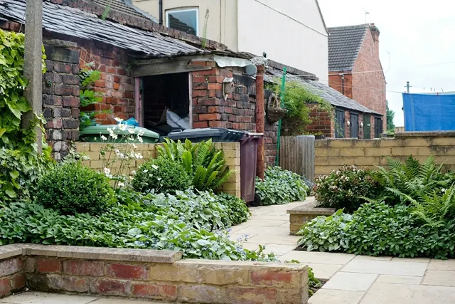 AI generated shade planting redesign of the same view: a dilapidated backyard with old brick outbuildings, a low wall, rubble, a discarded sink and weeds, replanted.