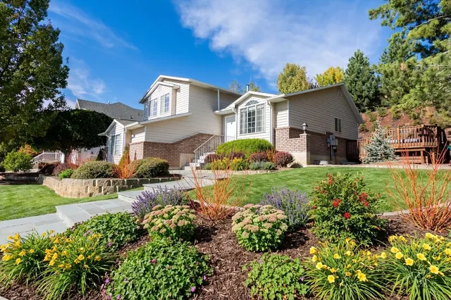 AI generated low maintenance redesign of the same view: a sloping lawn in front of a split-level house with a low stone retaining wall and shrub beds, replanted.