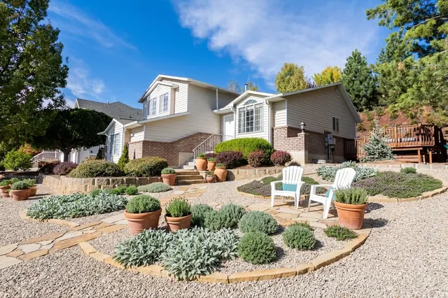 AI generated mediterranean redesign of the same view: a sloping lawn in front of a split-level house with a low stone retaining wall and shrub beds, replanted.